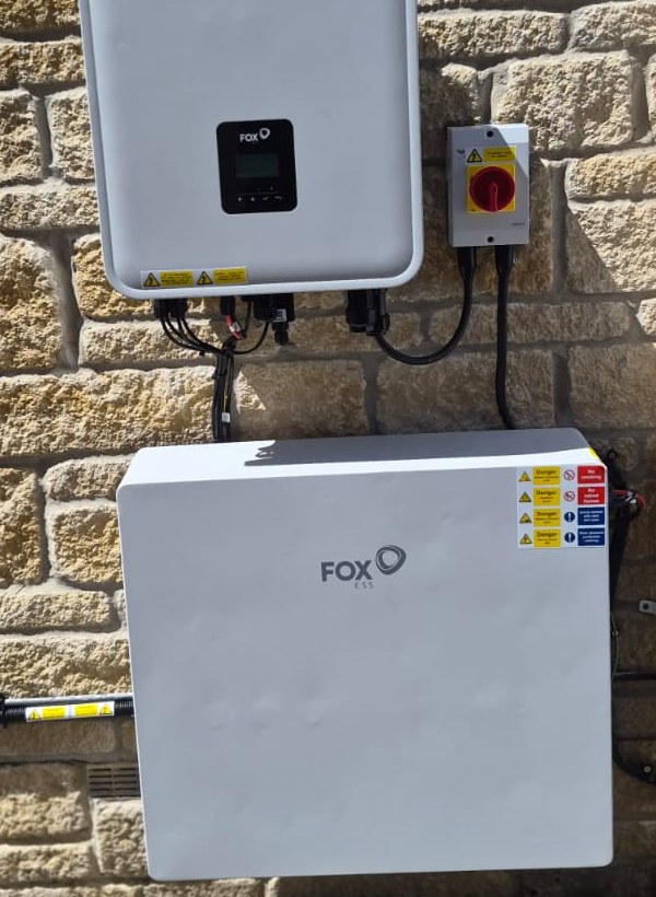 Fox ESS inverter with a red rotary isolator switch and a wall-mounted battery unit fixed to an exterior stone wall.