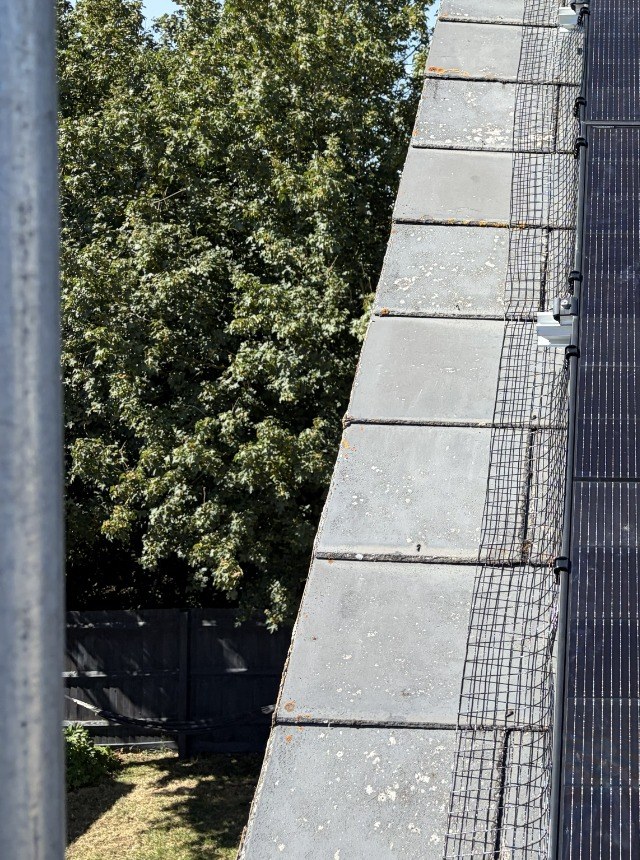Close view of the panel edge with black bird mesh fitted along the verge of a tiled roof, beside a stone chimney.