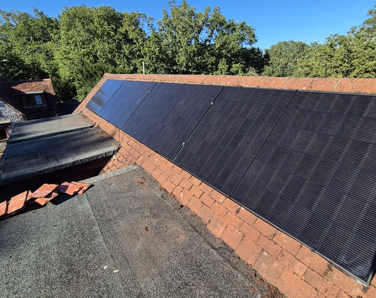 A long row of all-black solar panels fitted along a brick parapet roof under a clear blue sky, photographed by Cozy Home Solutions.